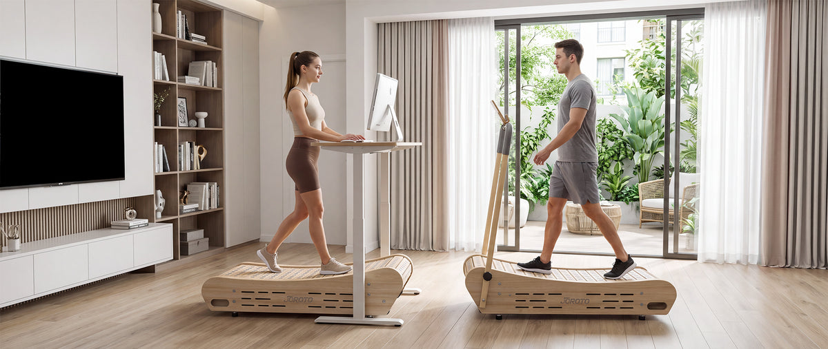 Two people exercise indoors on large wooden treadmills with built-in desks, set in a bright modern living room with large windows and curtains.